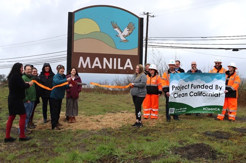 A small crowd of residents, Caltrans staff and local officials cut a ceremonial orange ribbon with large scissors in front of a gateway monument sign reading Manila with an osprey in midflight about to land on a stylized beach with a green hillside and sun rising in the background. 