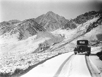 An undated black and white picture of a single vehicle driving on a snowy highway with snowy mountains in the distance.