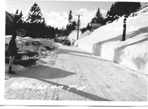 A black and white image of State Route 203 showing a man watching on as several cars follow a snowblower through Mammoth Lakes on January 1, 1966.