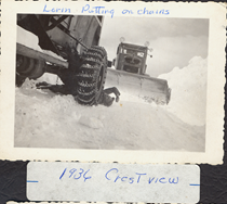 A black and white image of an unknown man putting snow chains on his vehicle with a snow plow behind him in 1936. Writing on the picture reads "Lorin Putting on chains. 1936 Crestview."